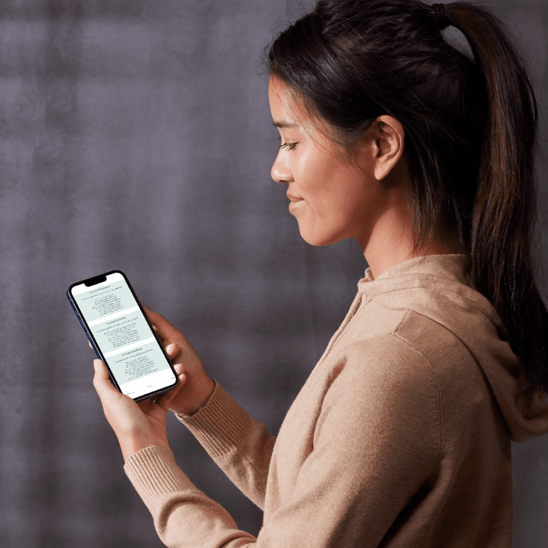 A woman in a tan hoodie stands in profile, holding and reading the "Trauma-Informed Alternatives to 'I Love and Accept Myself' – EFT Setup Phrase Guide (Printable PDF)" on her smartphone against a gray background.