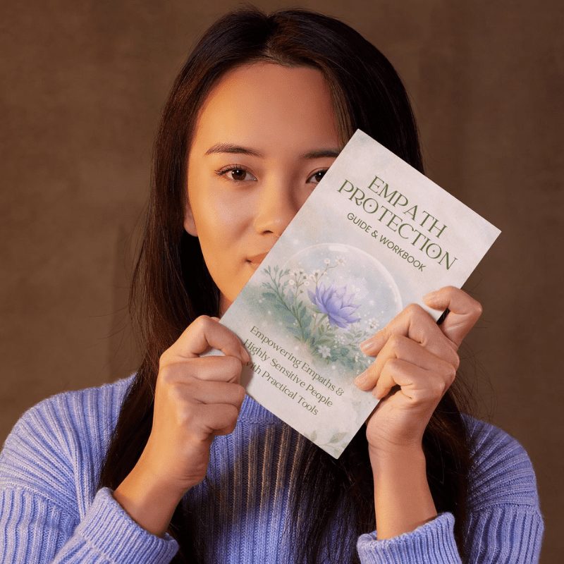 A woman in a lavender sweater holds up the "Empath Protection Guide & Workbook – Boundaries & Self-Care for Empaths & HSPs," partially covering her mouth.
