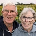 Two older adults smiling and posing for a selfie outdoors in front of a field of sunflowers.