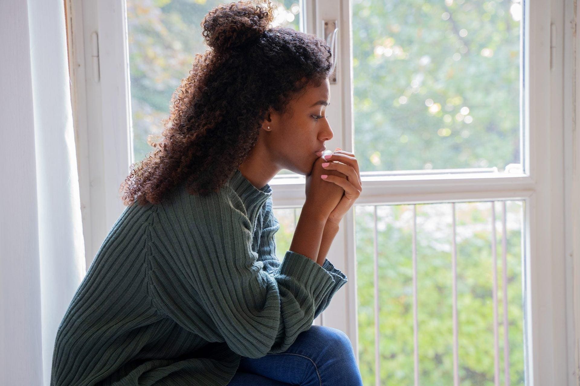 Young woman sitting indoors, looking thoughtfully out a window with her hands near her mouth, wearing a green sweater. Trees and a railing are visible outside.