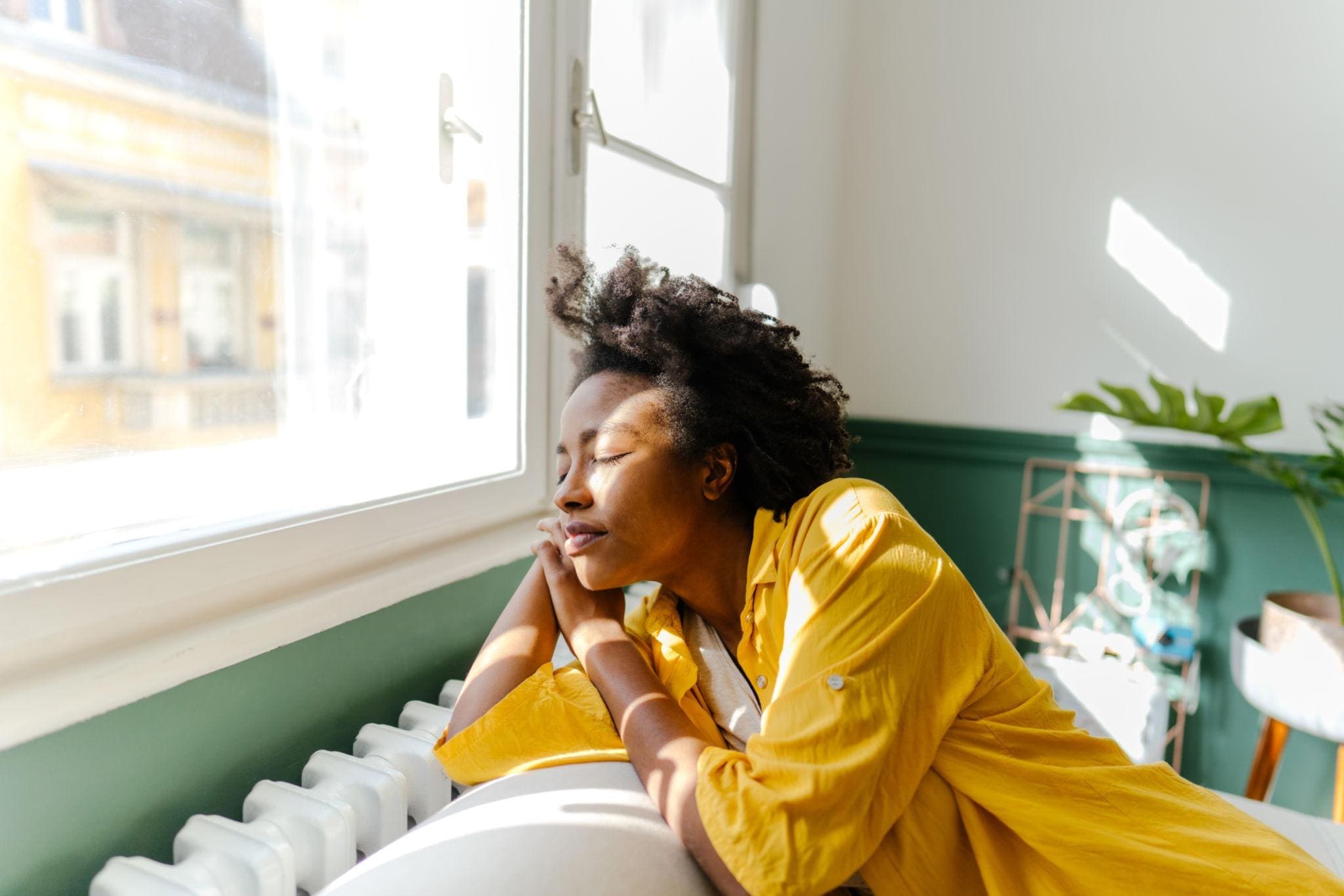 Person in a yellow shirt sits on a couch, resting their head on their hand and looking peaceful while sunlight streams through a nearby window.